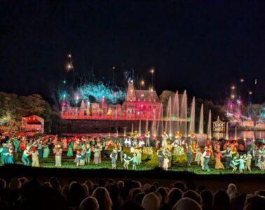 Puy du Fou at night with ruined huge house at back, lake and thousands of actors dancing in front dressed as peasants, audience in foreground