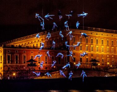 aquanauts show Caen Millennium with athletes suspended in mid air dressed in blue with building behind at night