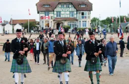 Canada House D-Day celebrations with band of bagpipers in Scottish tartans marching away from Canada House on beach