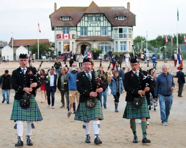 Canada House D-Day celebrations with band of bagpipers in Scottish tartans marching away from Canada House on beach