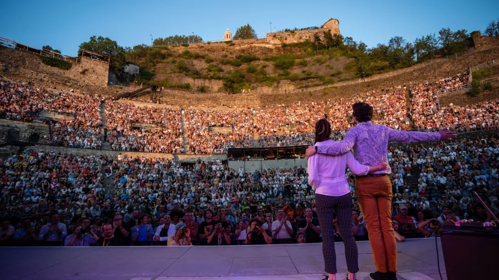 Orange amphitheatre with a couple in foreground on stage looking at crowds in background
