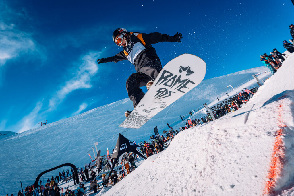 Rise Festival view from below snowboard jumpingon white board with Rise on bottom arms akimbo, snow and crowds