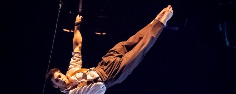Circus of Tomorrow man attached by feet and hand swinging through the airon trapeze