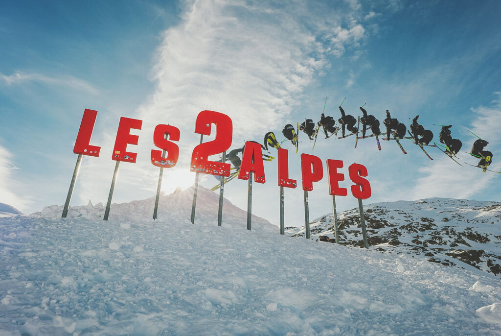 Les Deux Alpes red sign on snowy mountain with skiers jumping over it