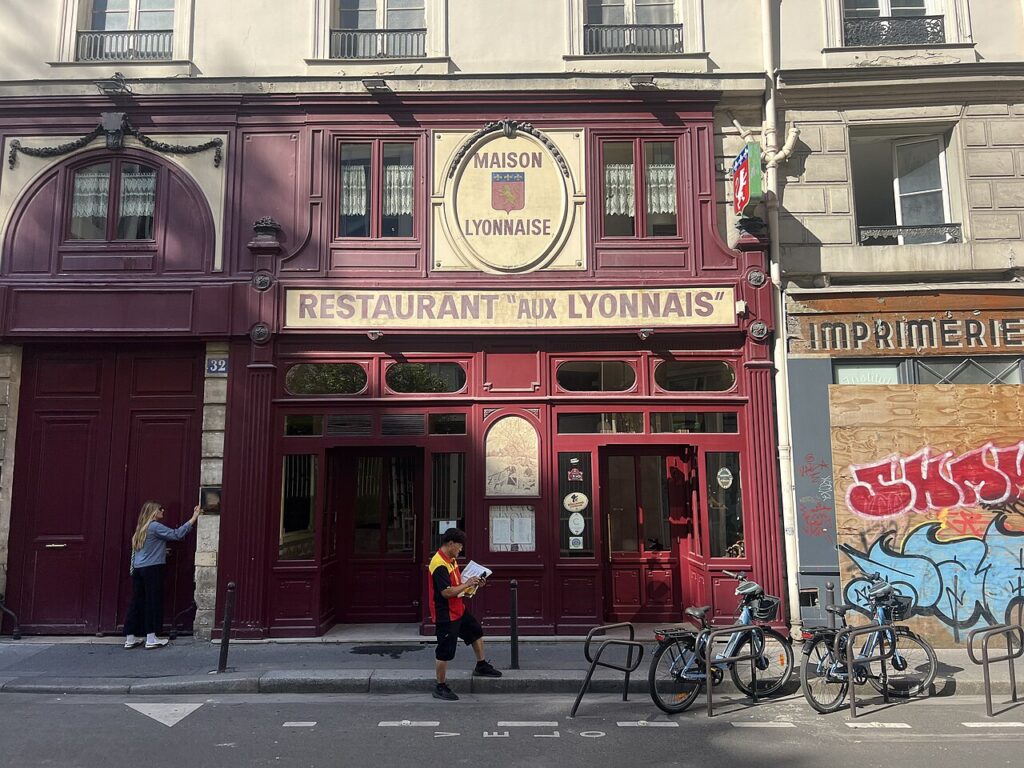Facade of Aux Lyonnais restaurant with wooden facade, awning with name and people passing by