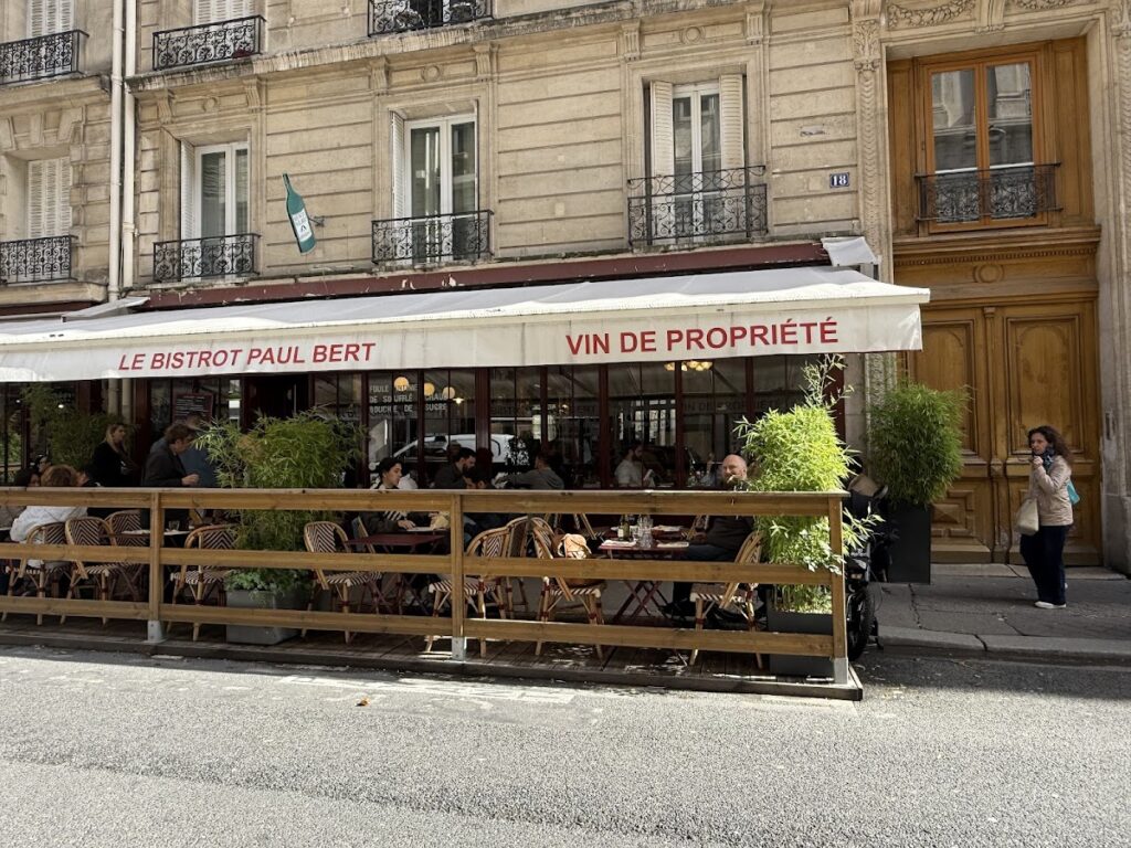 Le Bistrot Paul Bert Paris facade in old building with terrace with wooden fence, greenery and awning with name