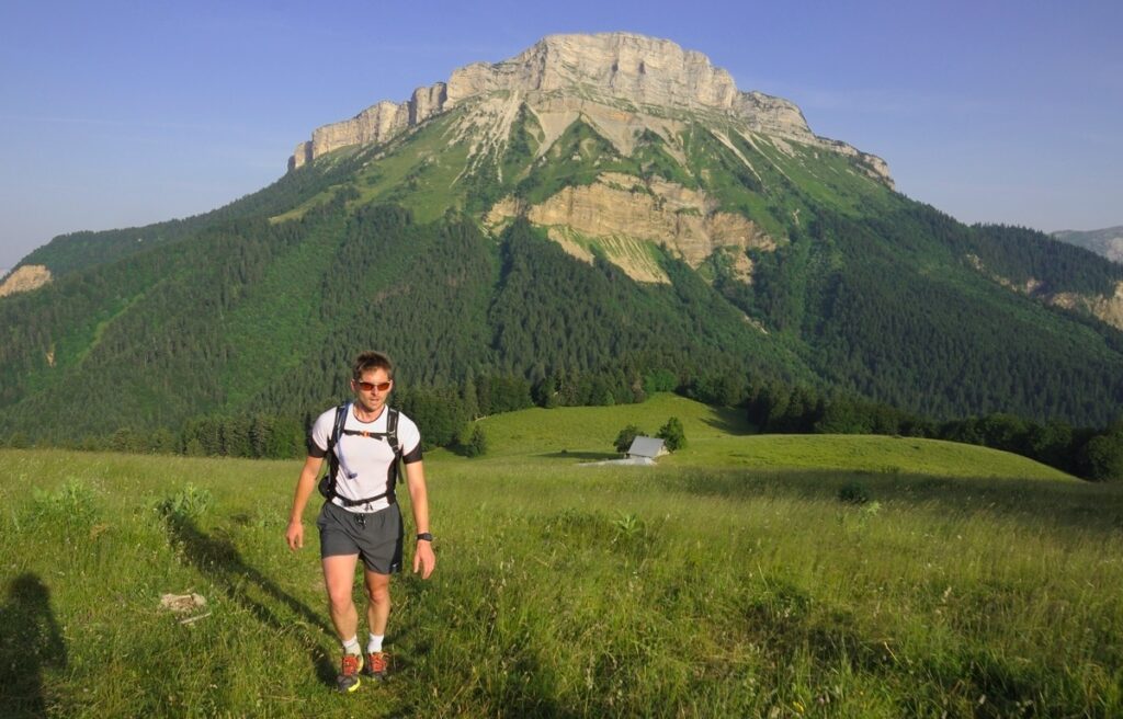 Solo man hiking in the Chartreuse in a field with a big bare mountain behind hime